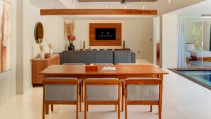 A modern dining area with a wooden table and chairs, a grey sofa, and a mounted television. - One Eleven Haven (Keroboken Kelod)