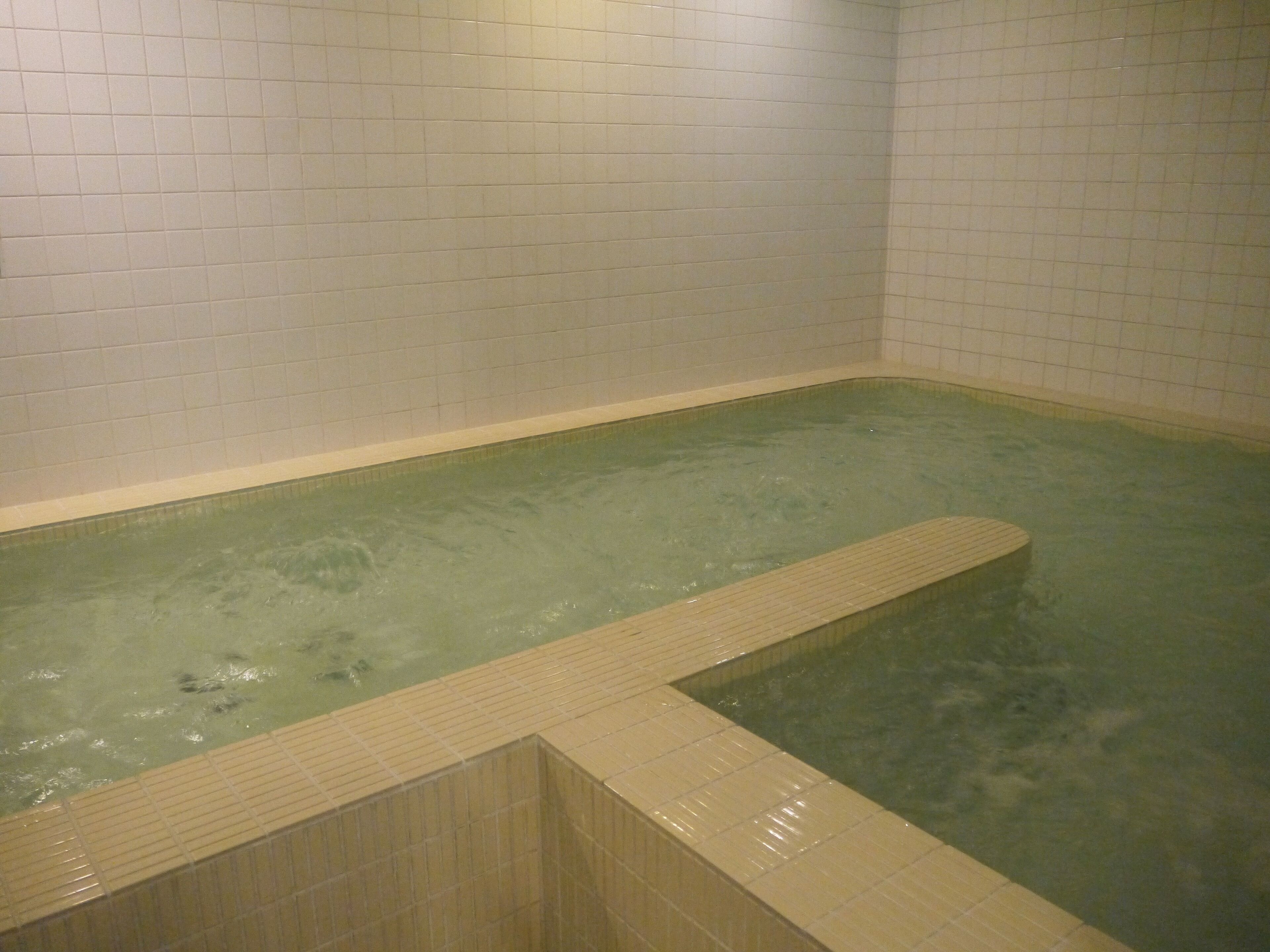 A small, rectangular hot tub with a wooden edge, surrounded by white tiled walls.