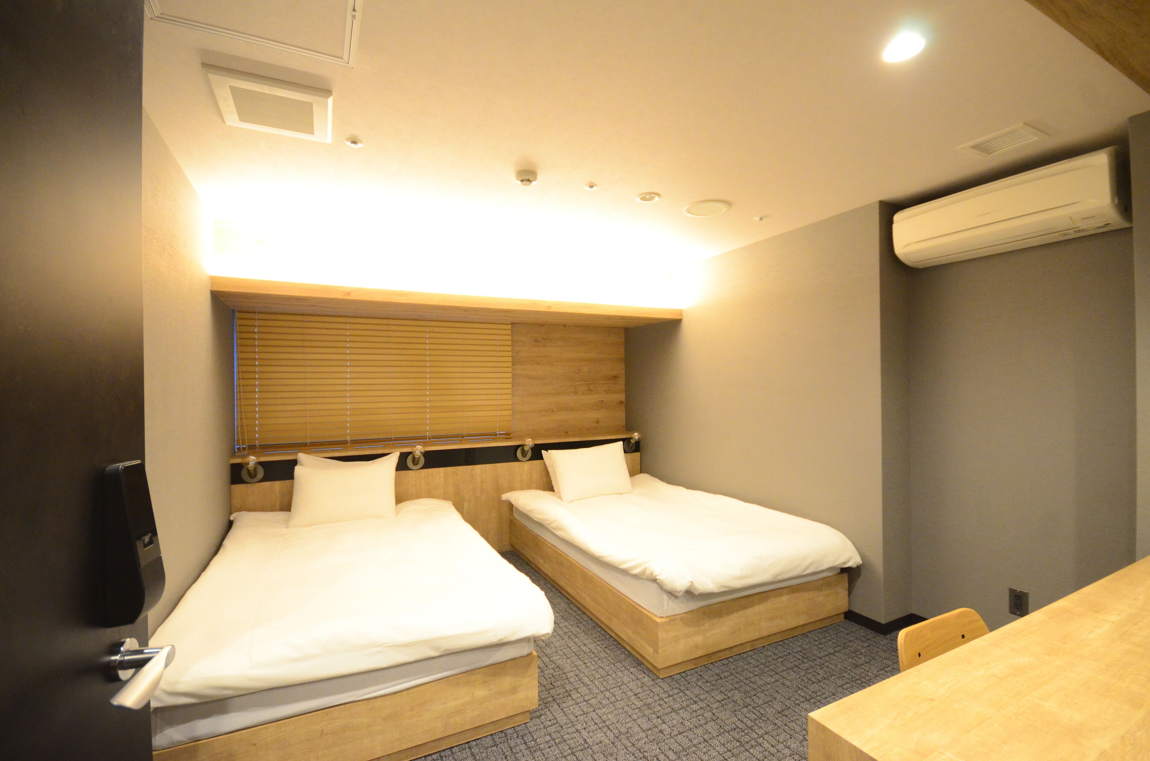 Habitación de hotel con dos camas individuales, cabecero de madera, unidad de aire acondicionado y una pequeña mesa.