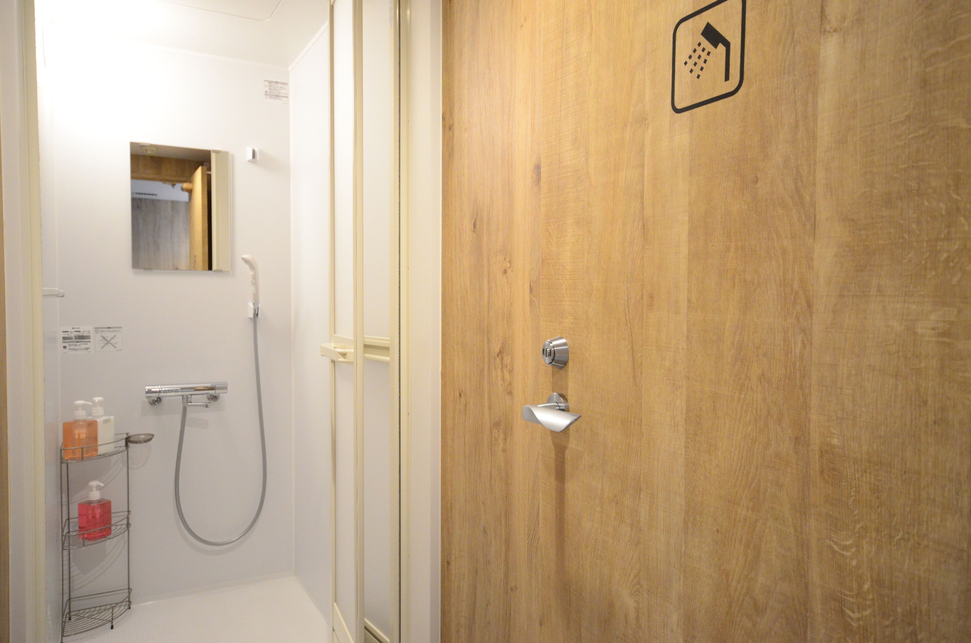 Un baño con puerta de madera, ducha, espejo y estante con artículos de aseo.