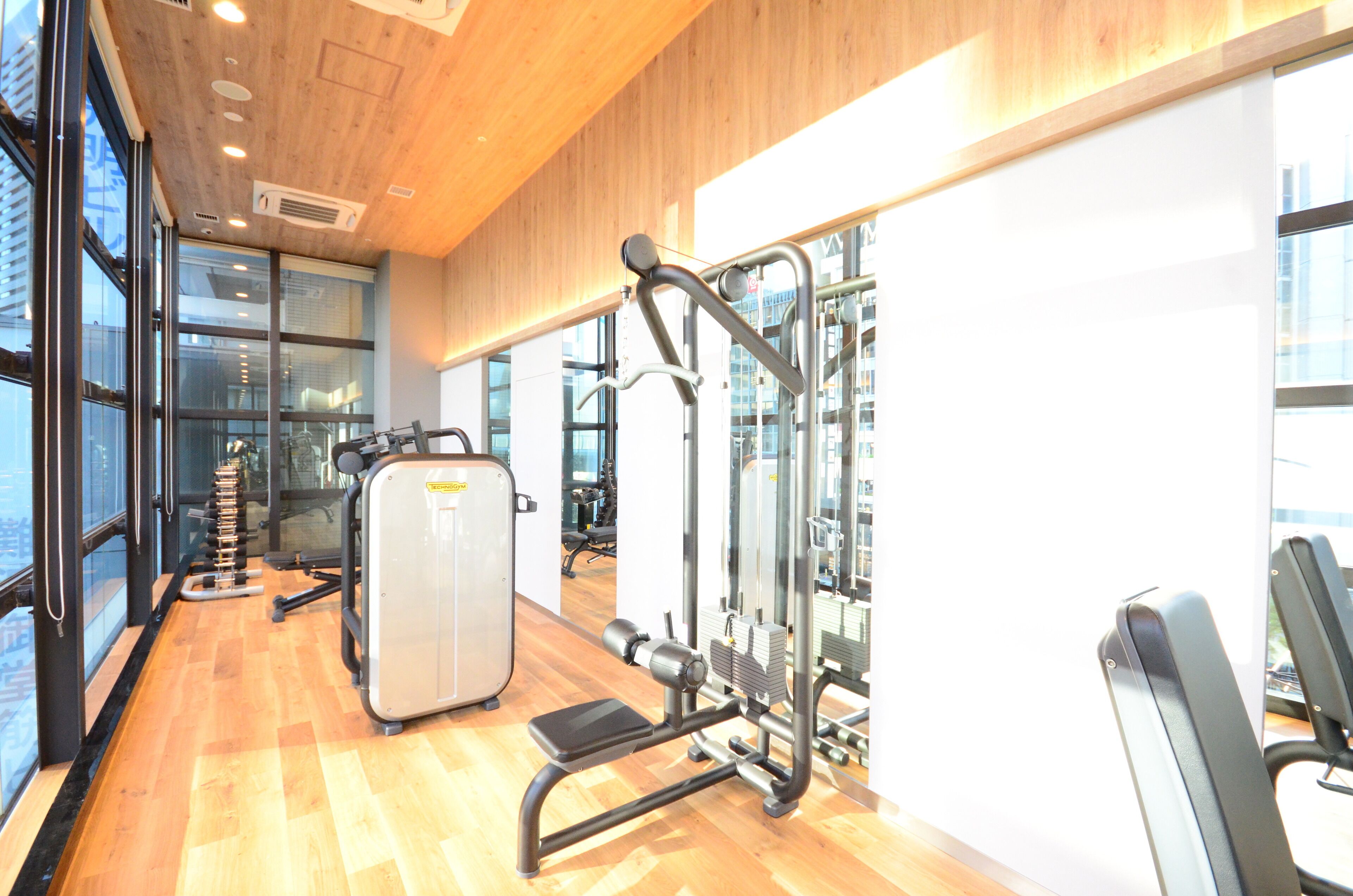 A gym with various exercise machines, large windows, and a wooden floor.