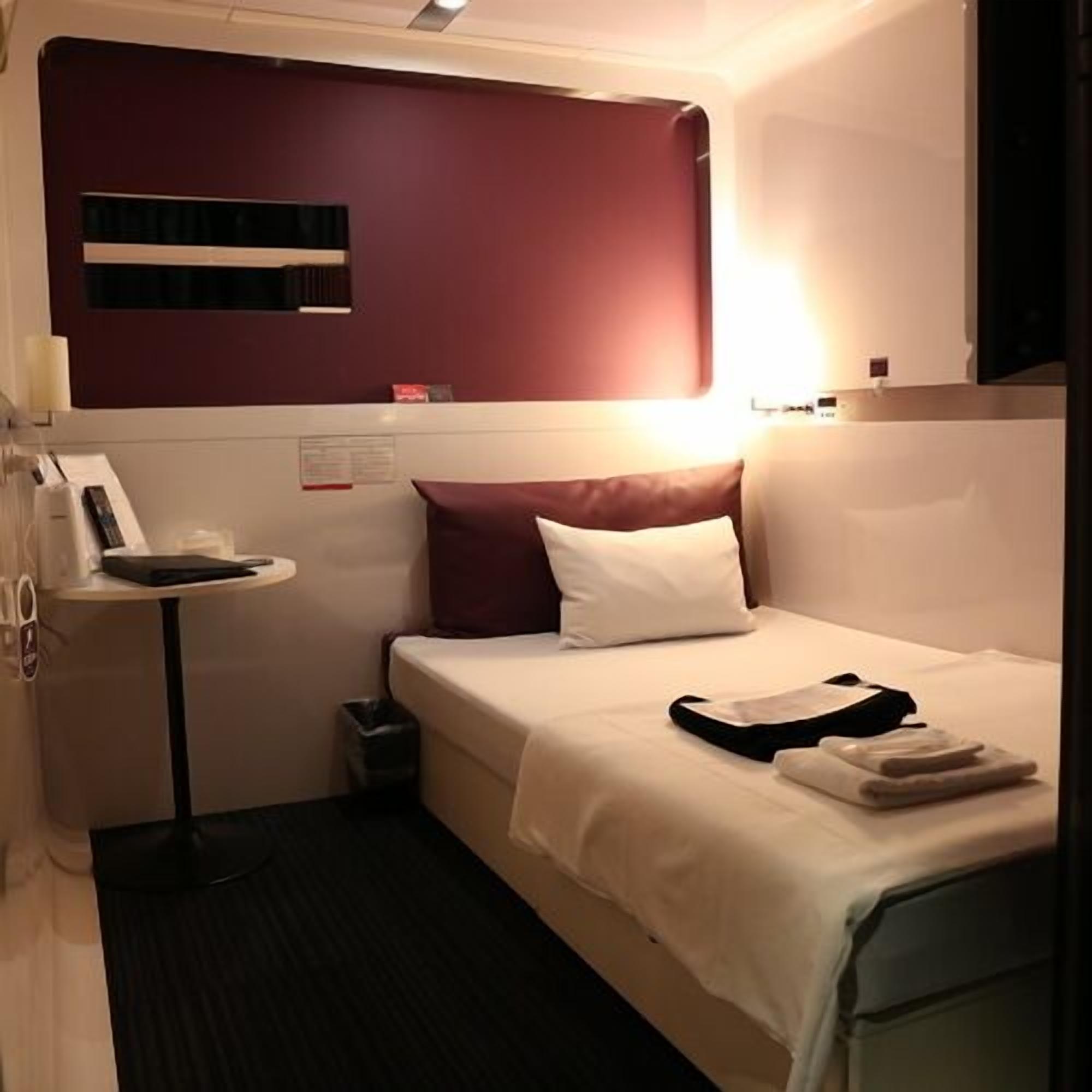 Une chambre d’hôtel comprenant un lit, une table de chevet, une lampe, une poubelle et une étagère fixée au mur.