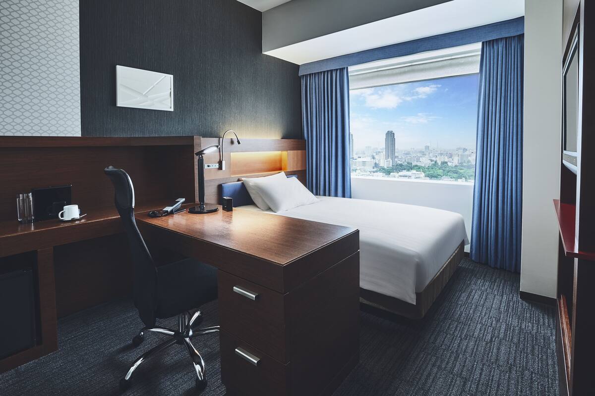 A hotel room with a bed, desk, chair, and a city view through large windows.