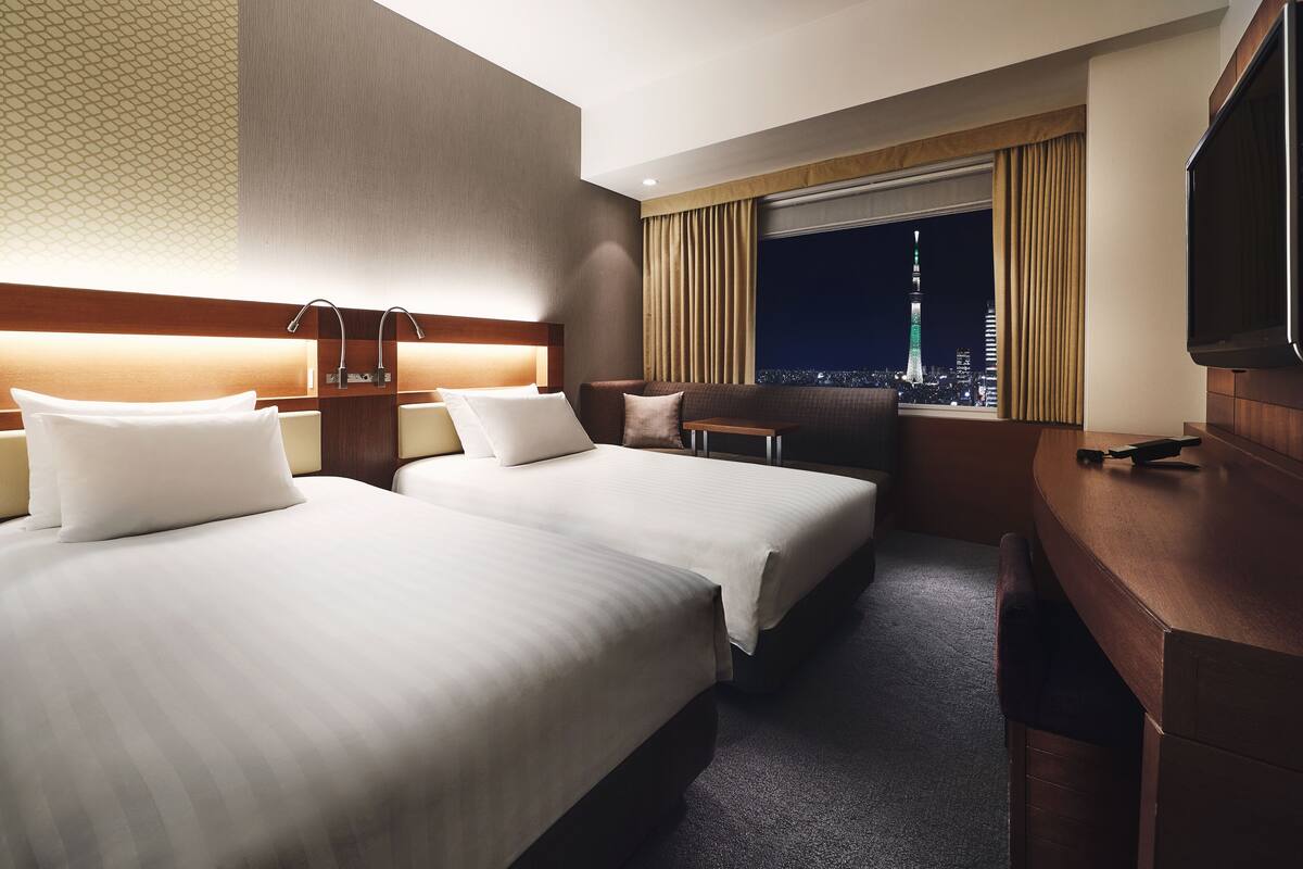 A hotel room with two beds, a desk, a TV, and a view of a cityscape at night.
