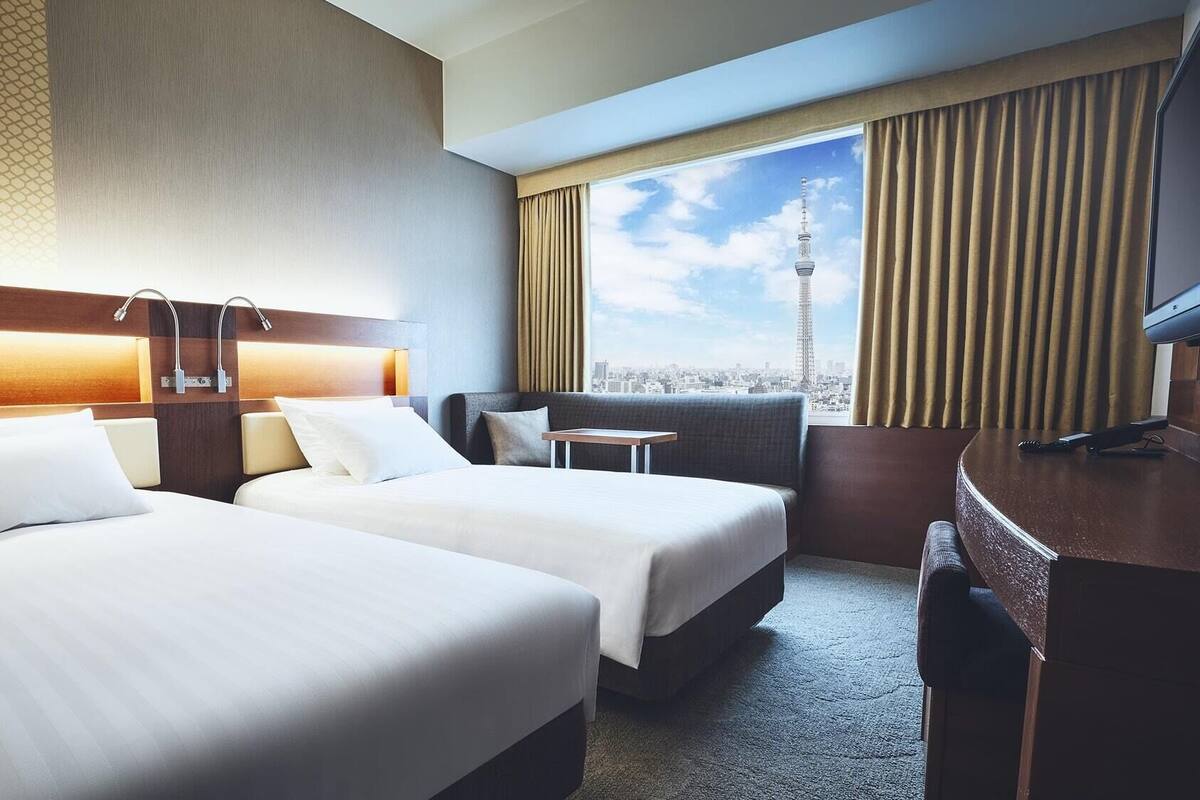 A hotel room with two beds, a desk, a chair, and a view of a cityscape including a tower.
