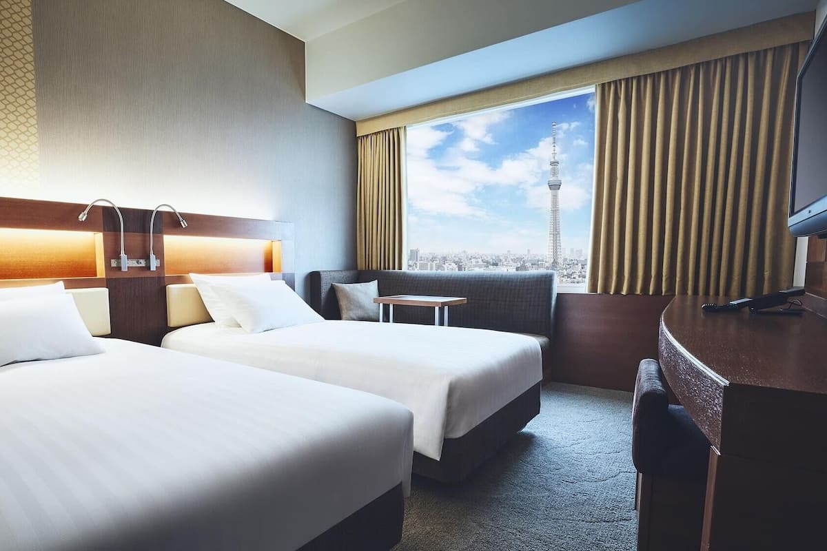 Une chambre d’hôtel avec deux lits, un bureau, une chaise et une vue sur le paysage urbain, notamment sur une tour.