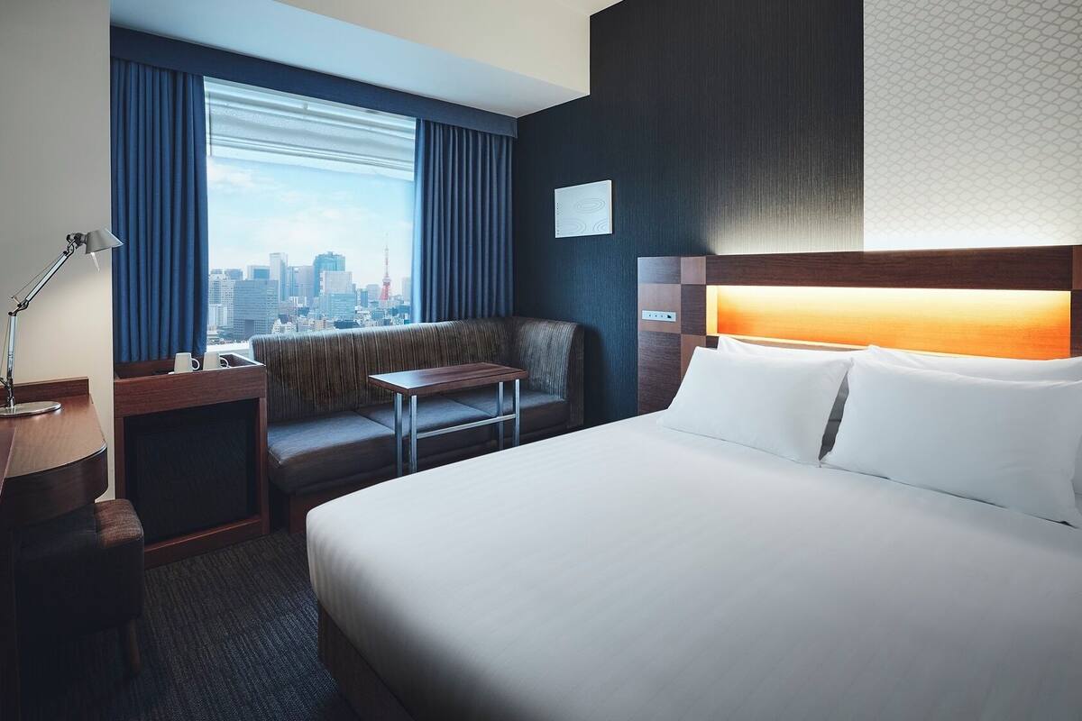 A hotel room with a bed, a desk with a lamp, a sofa, and a city view.