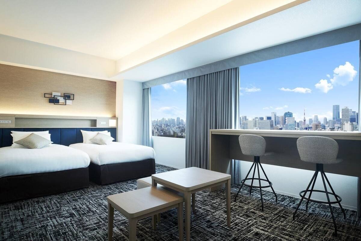 A hotel room with two beds, a small table, two bar stools, a city view, and a large window with curtains.