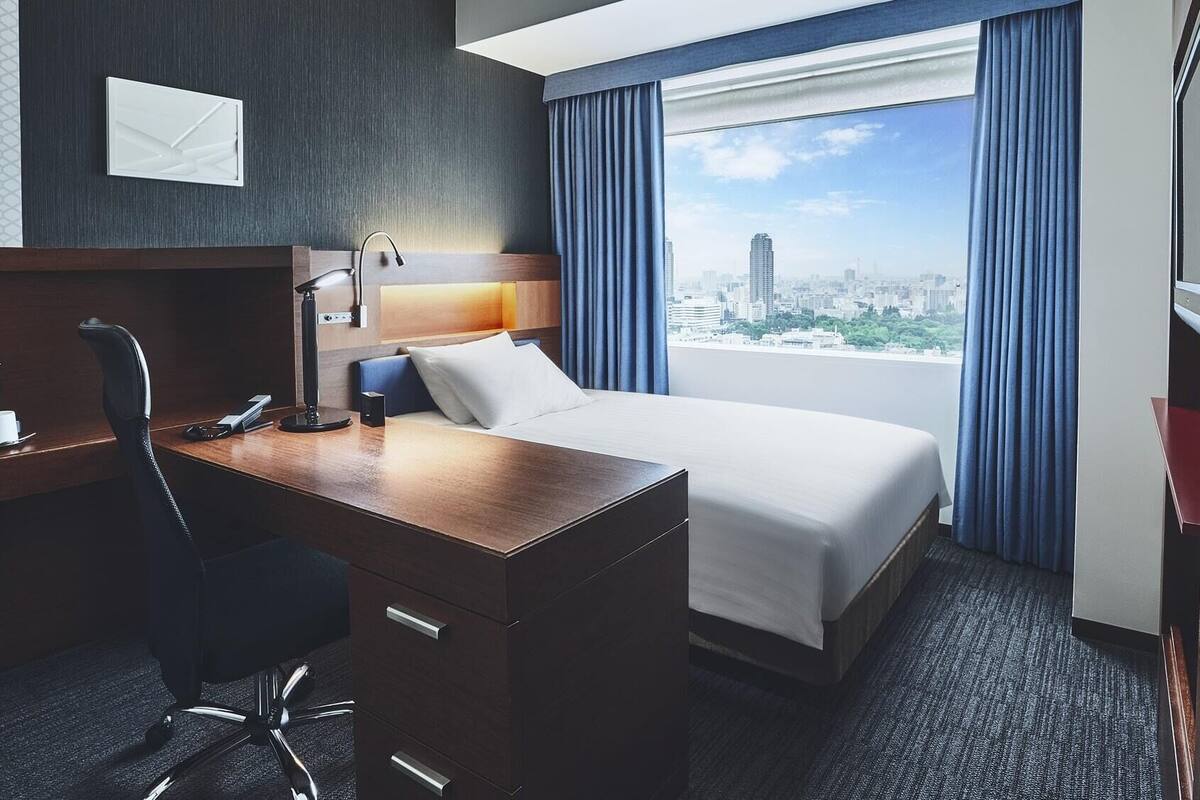 A hotel room with a bed, desk, chair, and a city view.