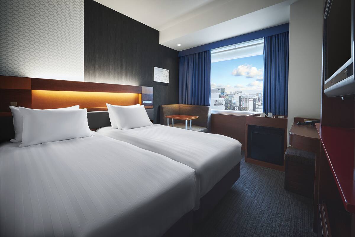 A hotel room with two beds, a desk, a chair, and a large window with a city view.
