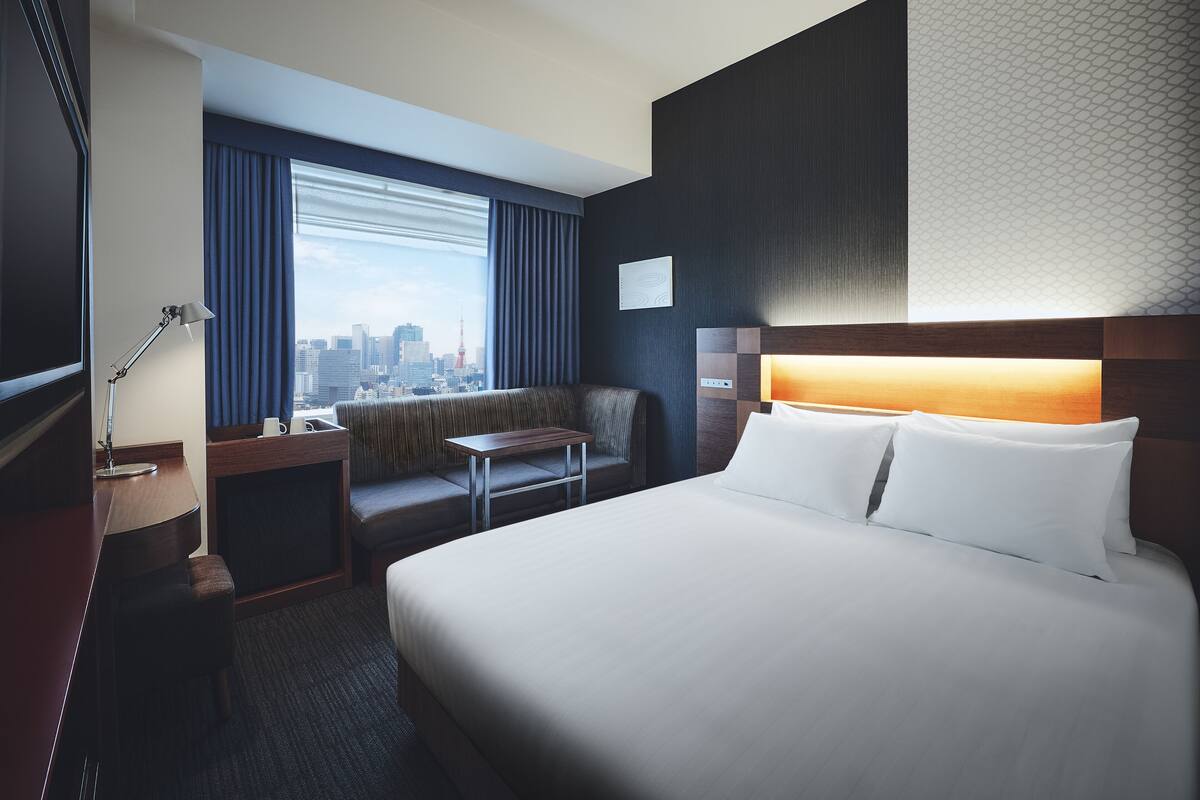 A hotel room with a large bed, a seating area with a view of the city, a desk with a lamp, and a TV.
