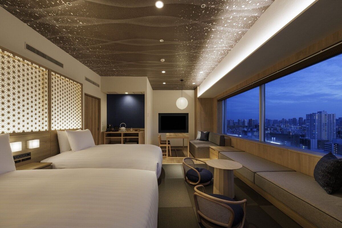 A hotel room with two beds, a TV, a desk, and a large window with a city view.