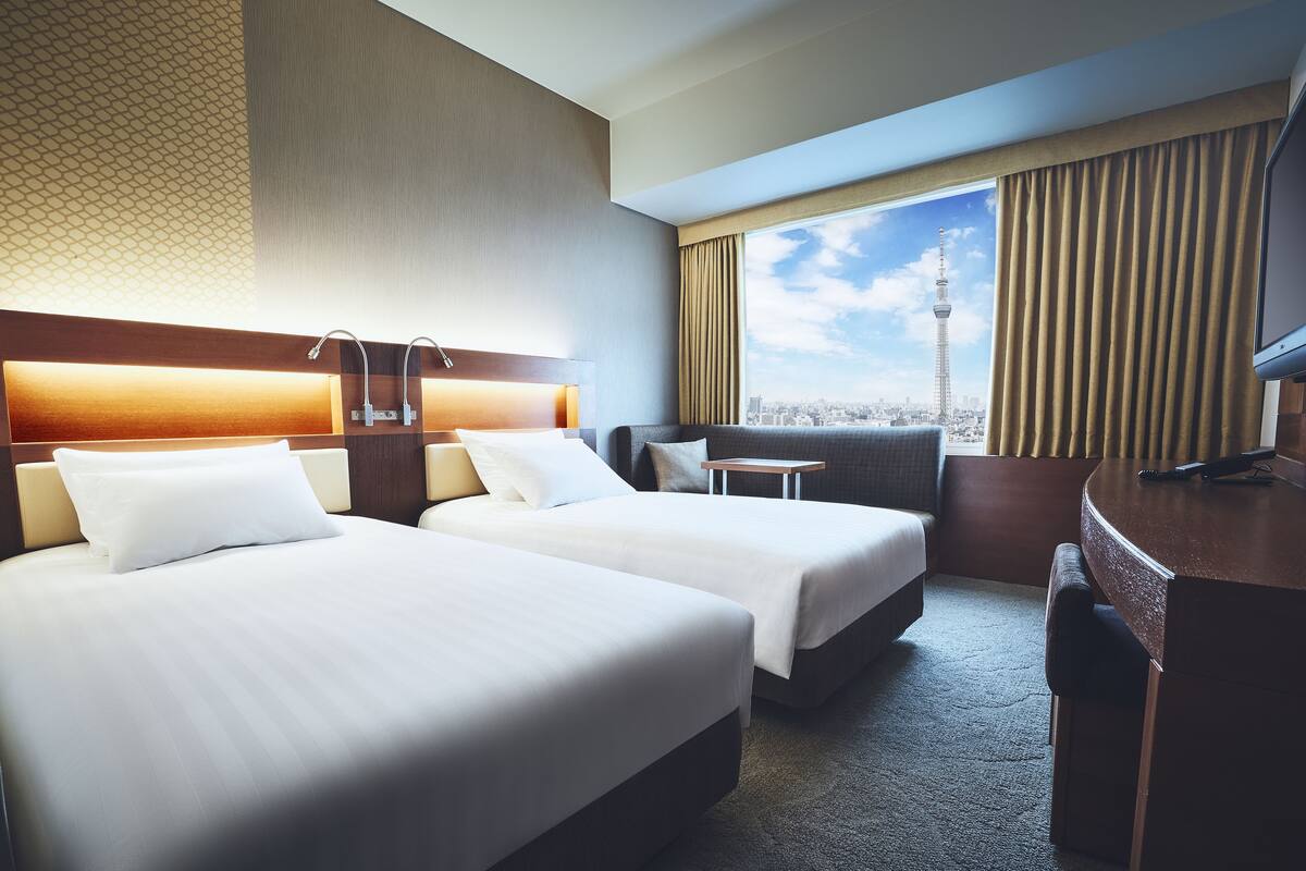 A hotel room with two beds, a desk, and a view of a cityscape including a tower.