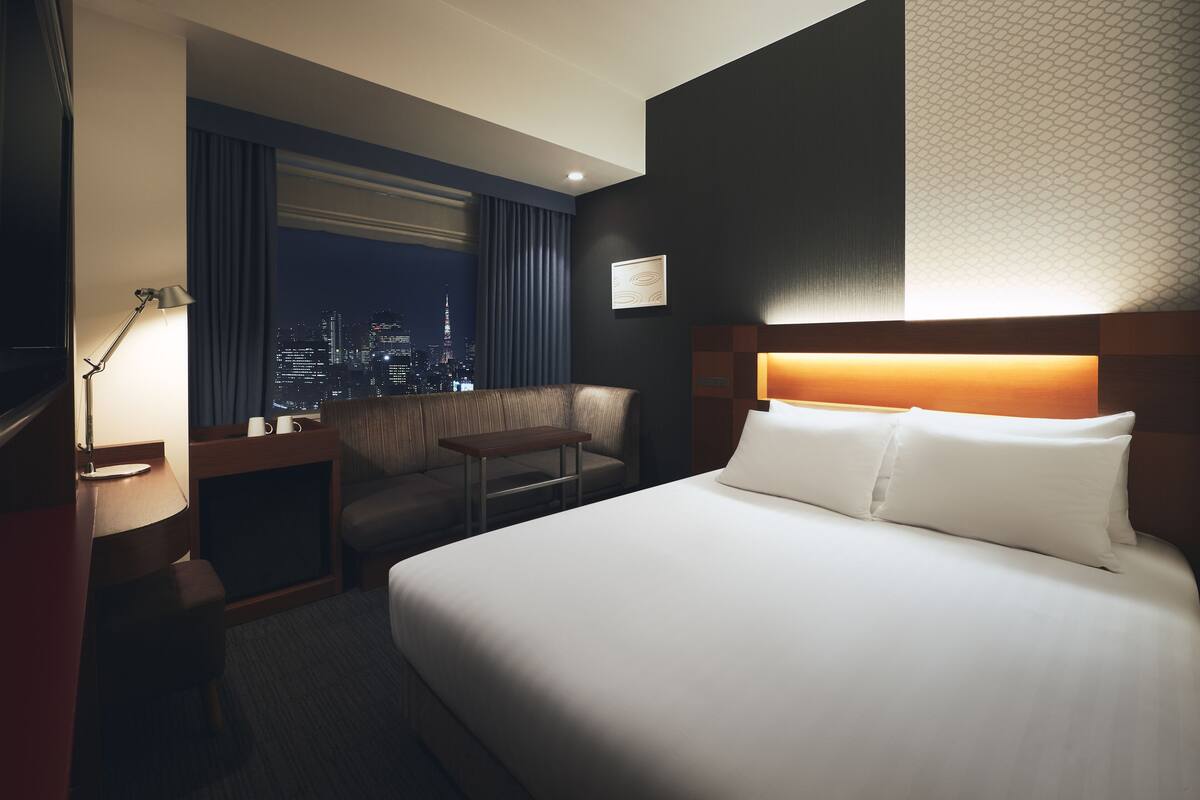 A hotel room with a large bed, a desk, a chair, and a view of a cityscape at night.