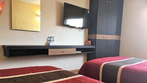 Une chambre d’hôtel avec deux lits, une télévision, une armoire et une étagère avec des bouteilles d’eau.