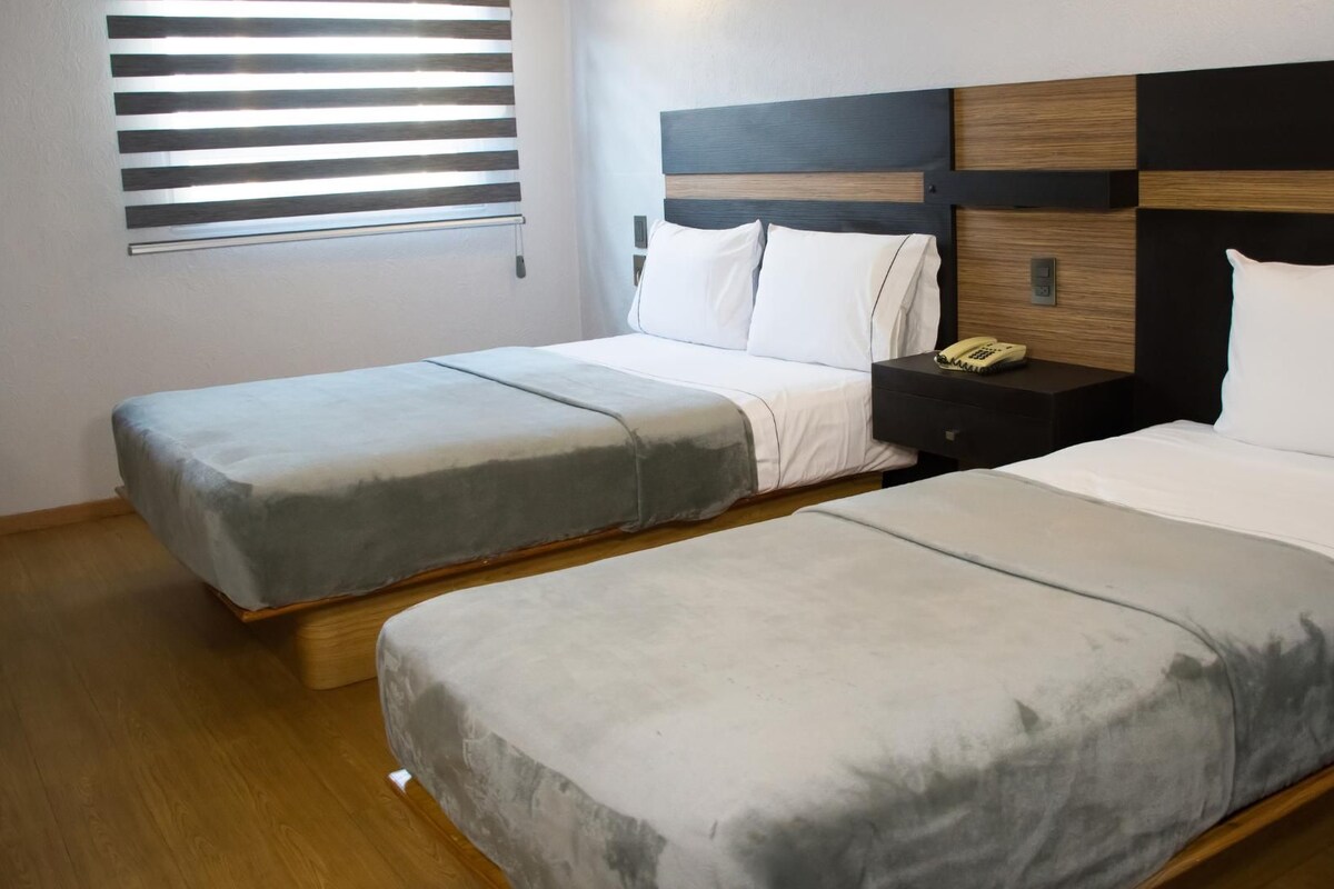 A hotel room with two single beds, a bedside table with a phone, and a window with blinds.