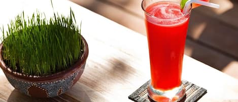 Un vaso de bebida roja con un adorno, sobre un posavasos, colocado sobre una mesa de madera junto a un cuenco decorativo con hierba verde.