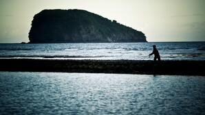 A coastal scene with a person walking along the shore, a large island in the background, and calm sea. - D Tunjung Resort & Spa (Karangasem)