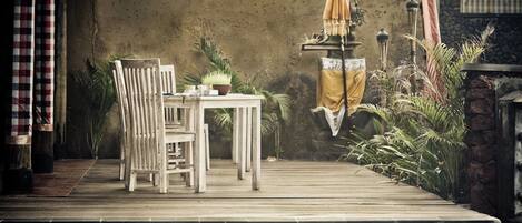 Une terrasse en bois avec une table et des chaises, une petite table ornée d’une plante et un parasol jaune.