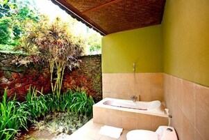Un baño con bañera, inodoro y ducha. Se aprecia una pared de piedra y vegetación en el exterior.