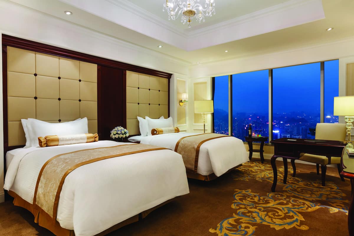 Ein Hotelzimmer mit zwei Betten, einem Schreibtisch und Blick auf die beleuchtete Stadt.
