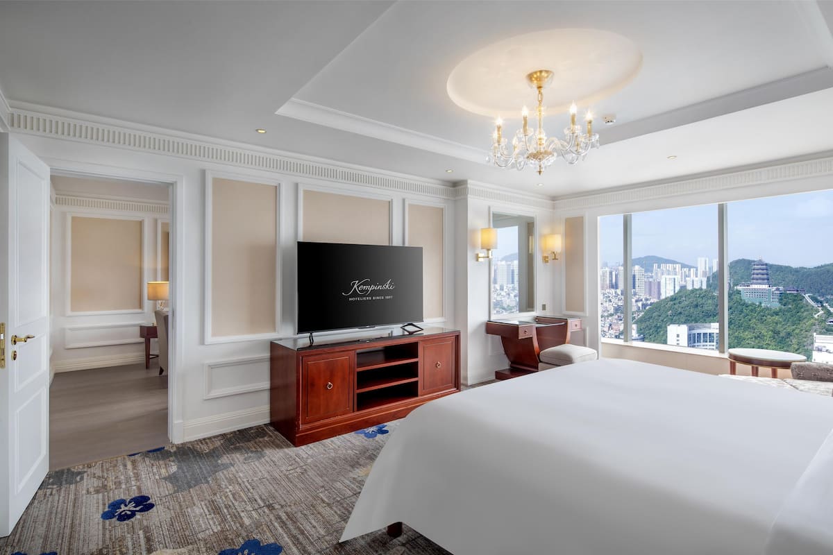 Ein Hotelzimmer mit einem großen Bett, einem Fernseher auf einer Holzkabine, Blick auf die Stadt und einer Kristallleuchter.