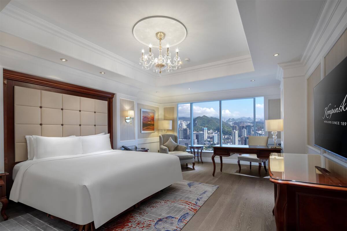 Ein Hotelzimmer mit einem großen Bett, einem Schreibtisch, einem Sessel und einem Blick auf die Stadt.