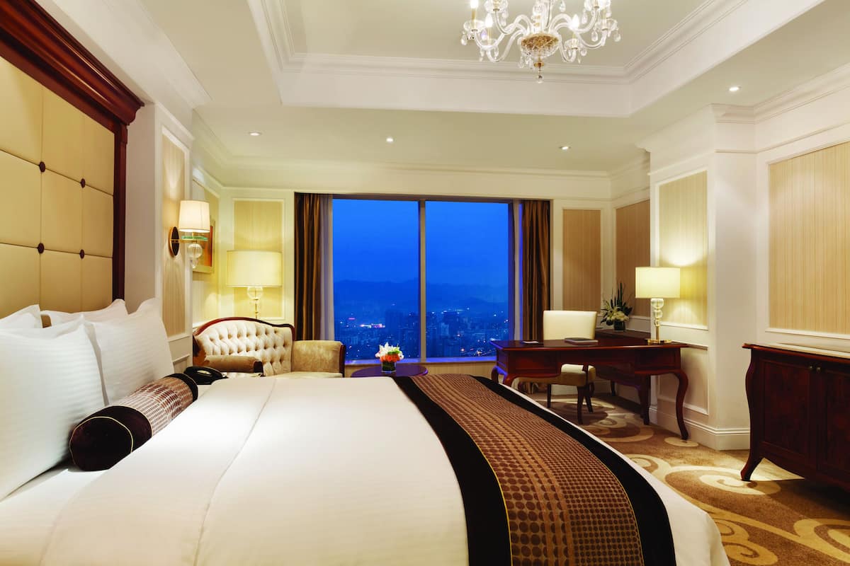 Ein geräumiges Hotelzimmer mit einem großen Bett, einem Schreibtisch und Blick auf die Stadt bei Einbruch der Dunkelheit.