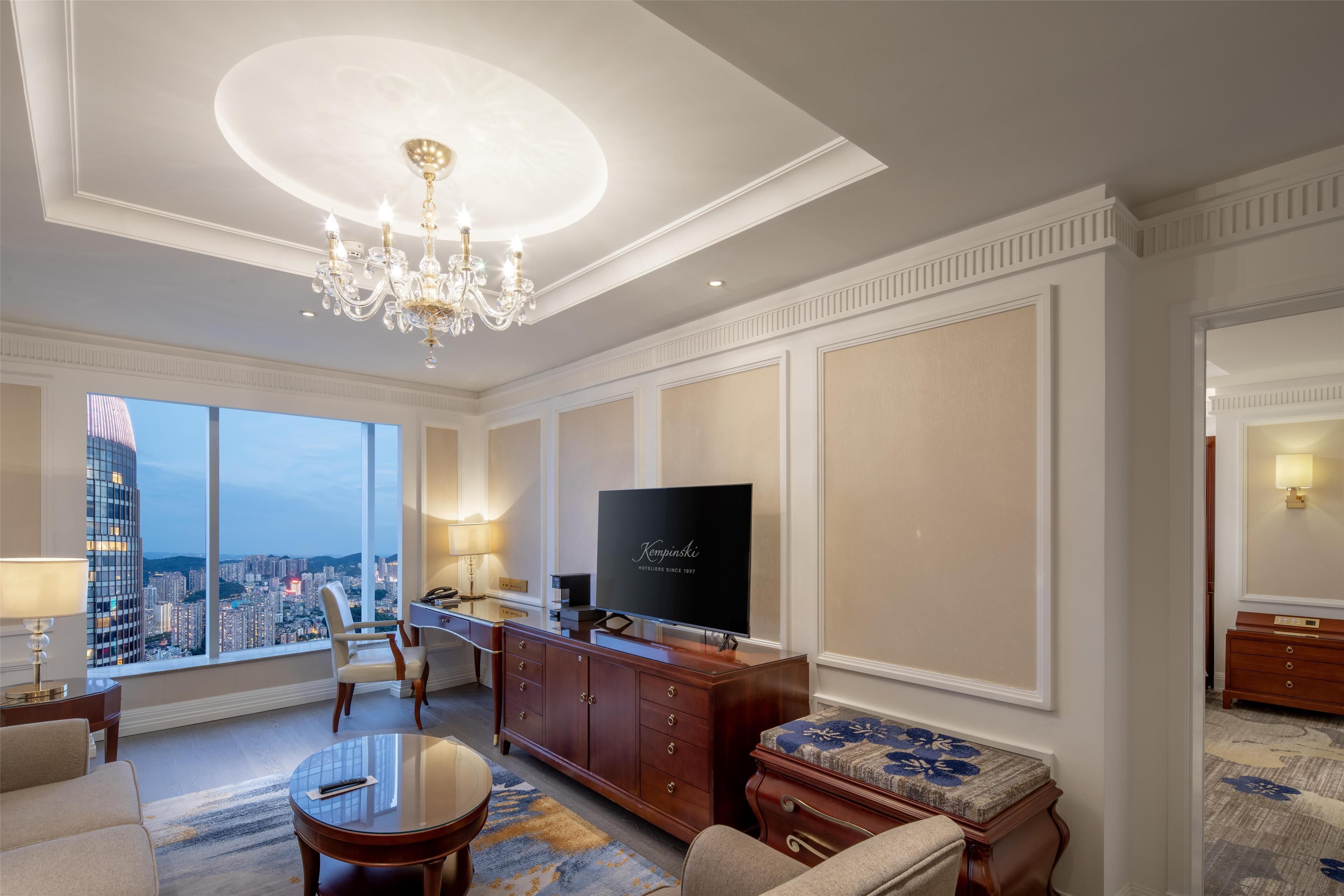 A spacious living room with a large window offering a city view, a chandelier, a flat-screen TV, and a comfortable seating area.
