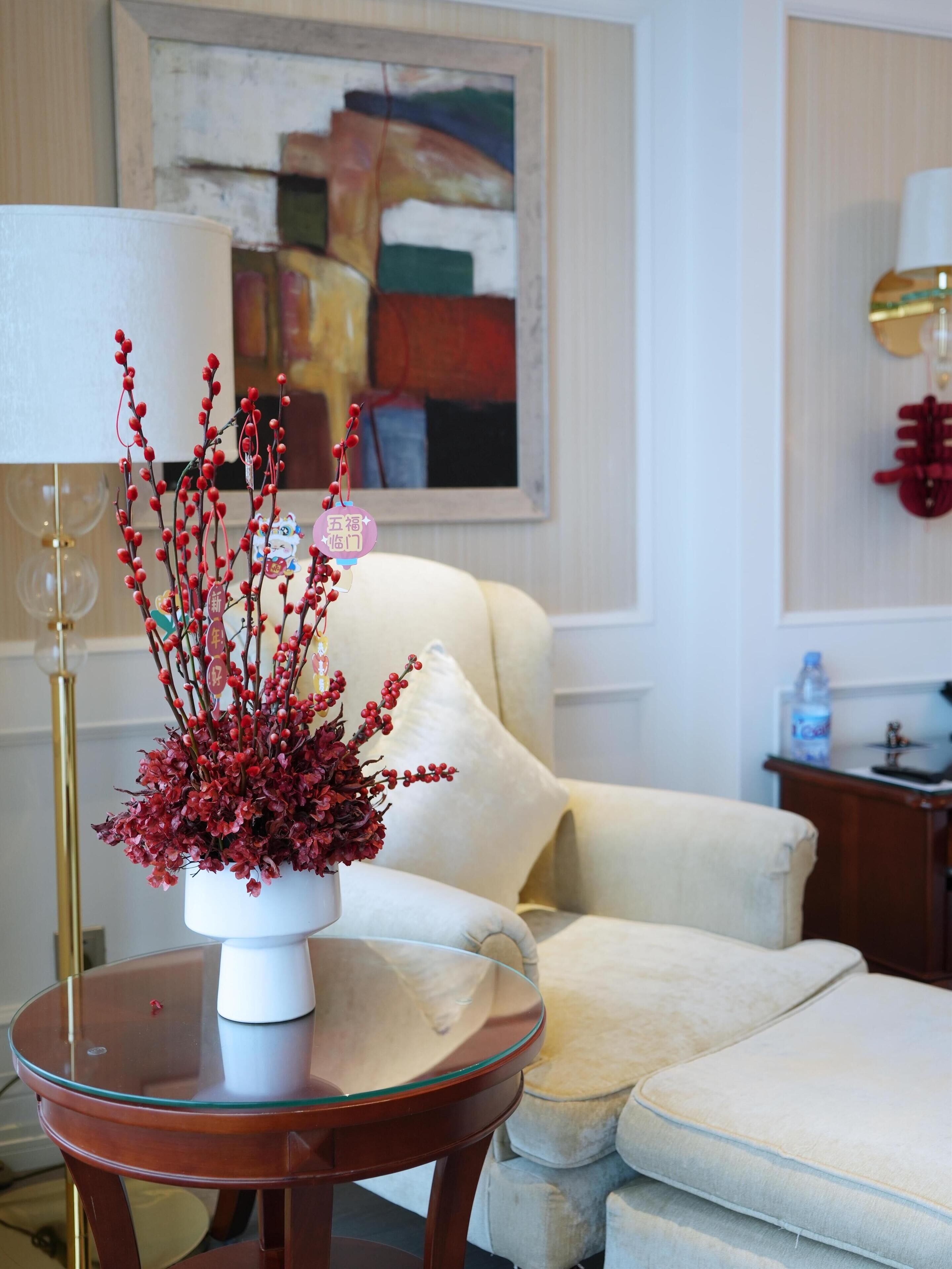 A living room with a white armchair, a glass table with a white vase containing red berries, a lamp, and a painting on the wall.