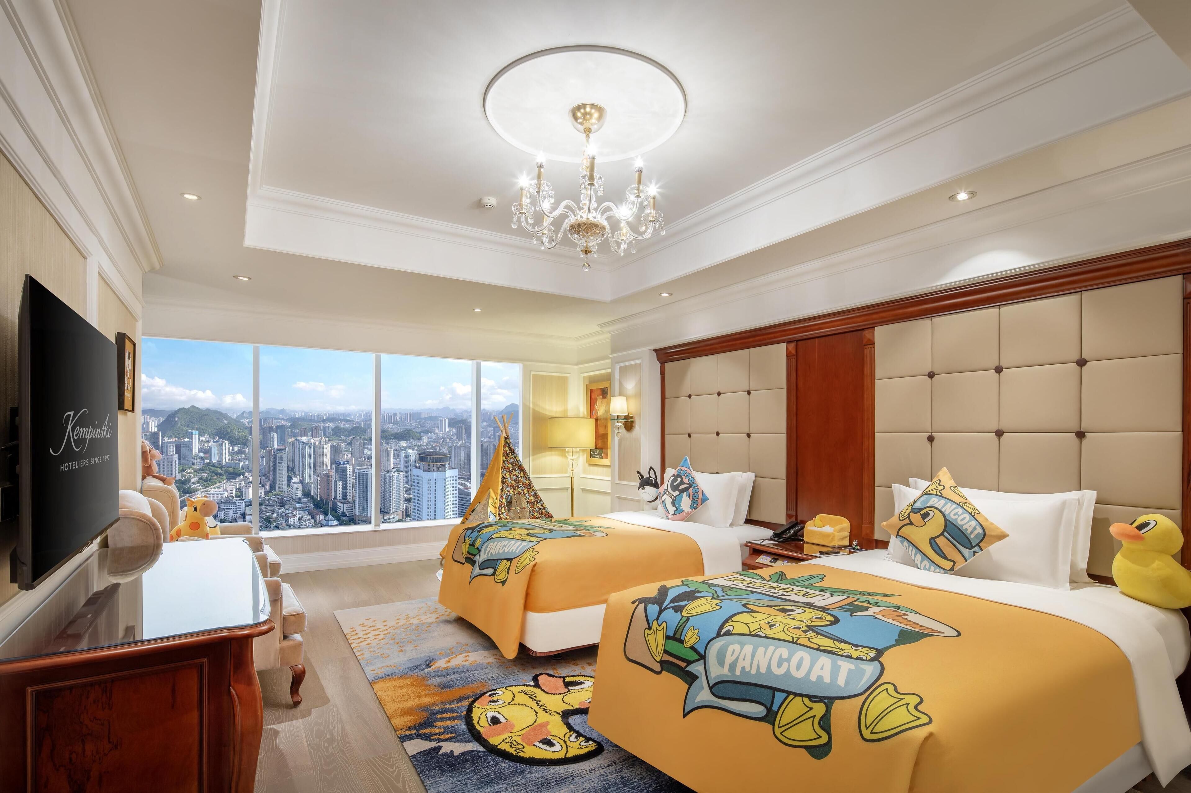 A hotel room with two beds, a large window with a city view, a chandelier, and a flat-screen TV.