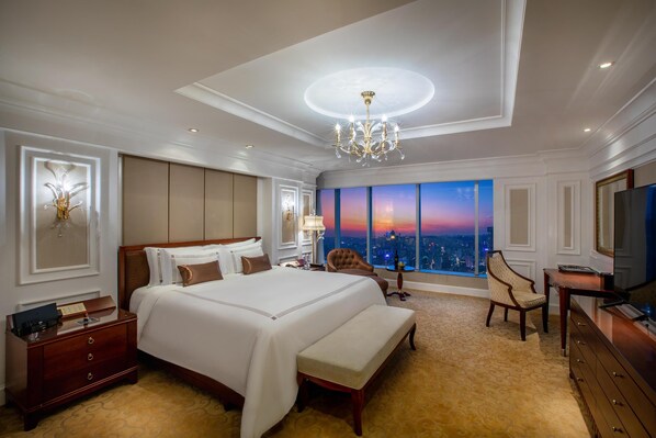 Amplia habitación de hotel con una cama grande, un banco, un televisor y vistas a la ciudad al atardecer.