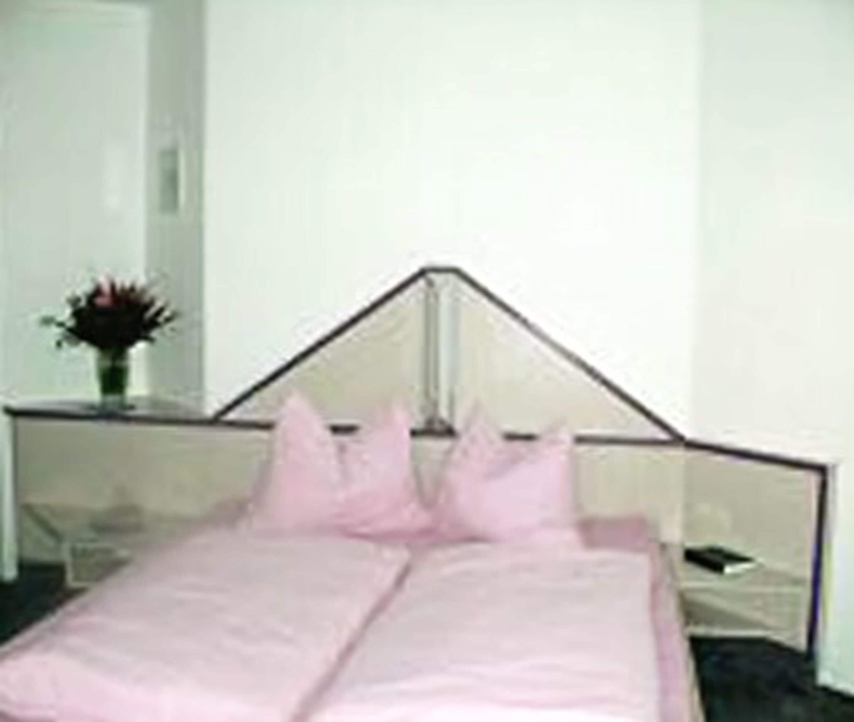Una cama con cabecera blanca y ropa de cama rosa en una habitación con paredes blancas.