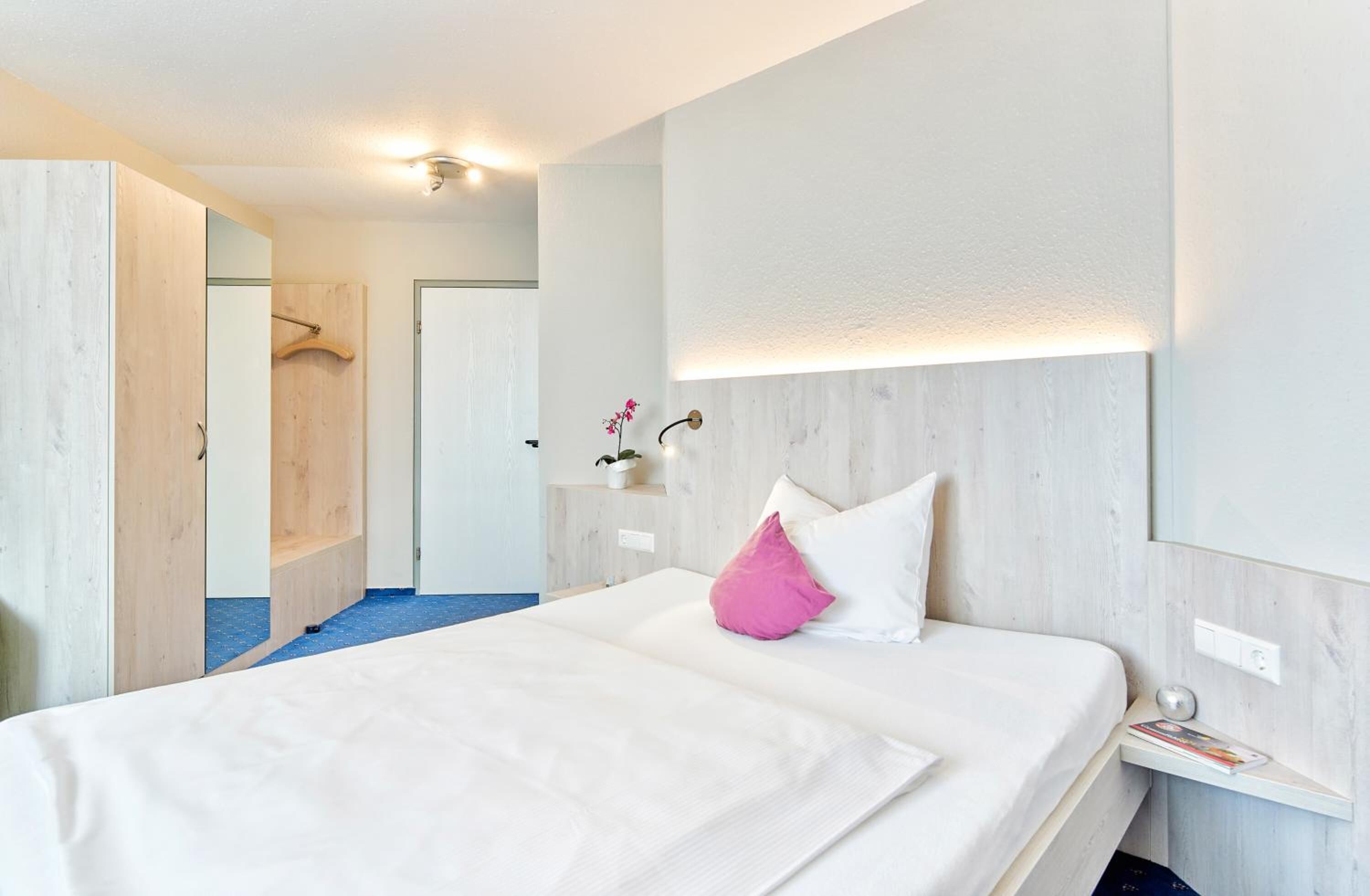 A hotel room with a large bed, a bedside table, a nightstand, a wall-mounted light, and a door leading to a bathroom.