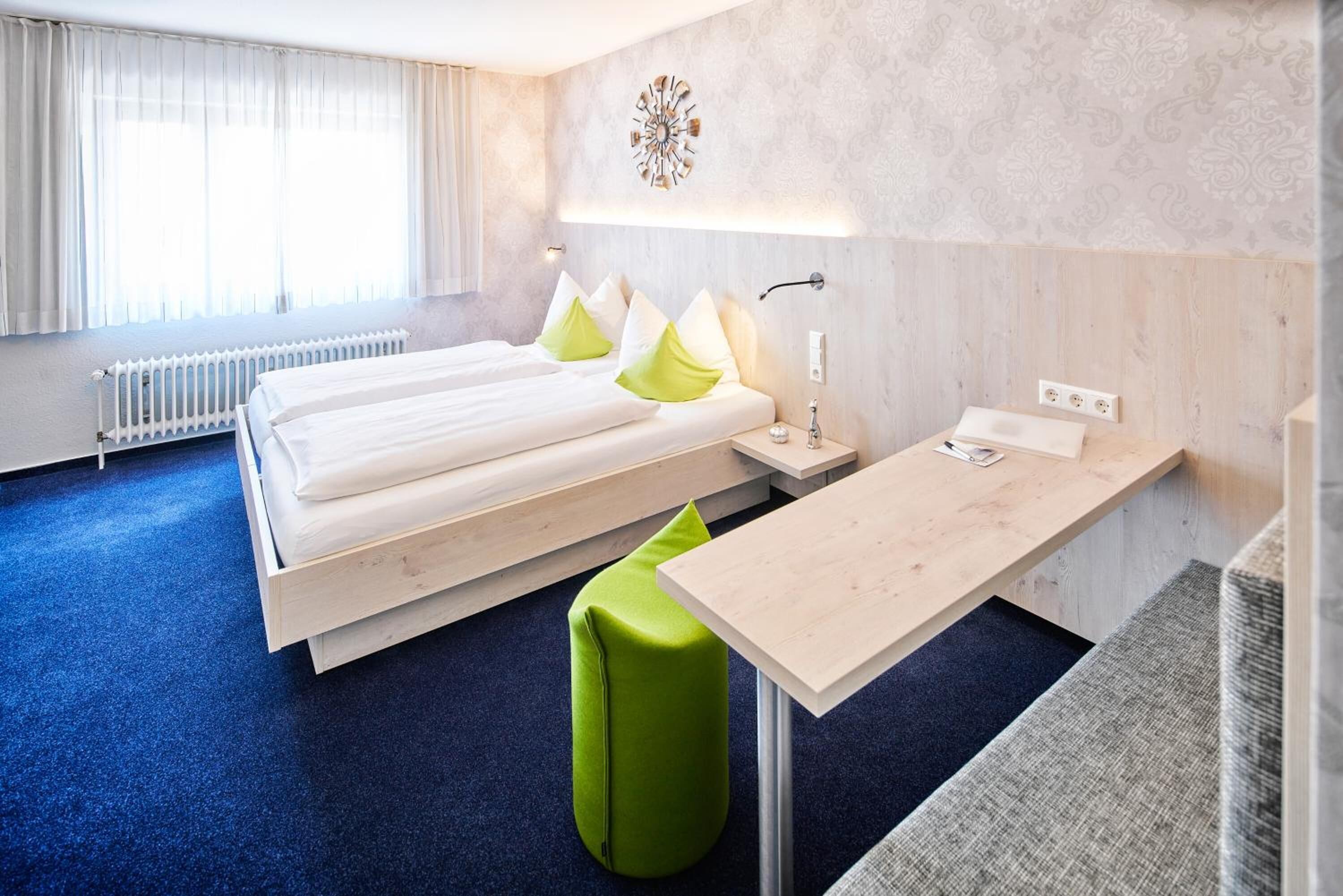 Ein Hotelzimmer mit zwei Betten, einem Schreibtisch, einem grünen Hocker und einer Wandleuchte.