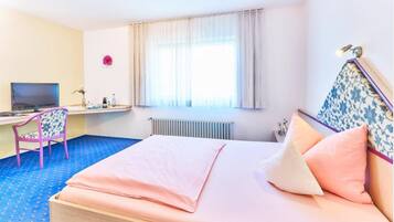 A hotel room with a bed, a desk with a computer, a chair, a radiator, and a window with curtains.