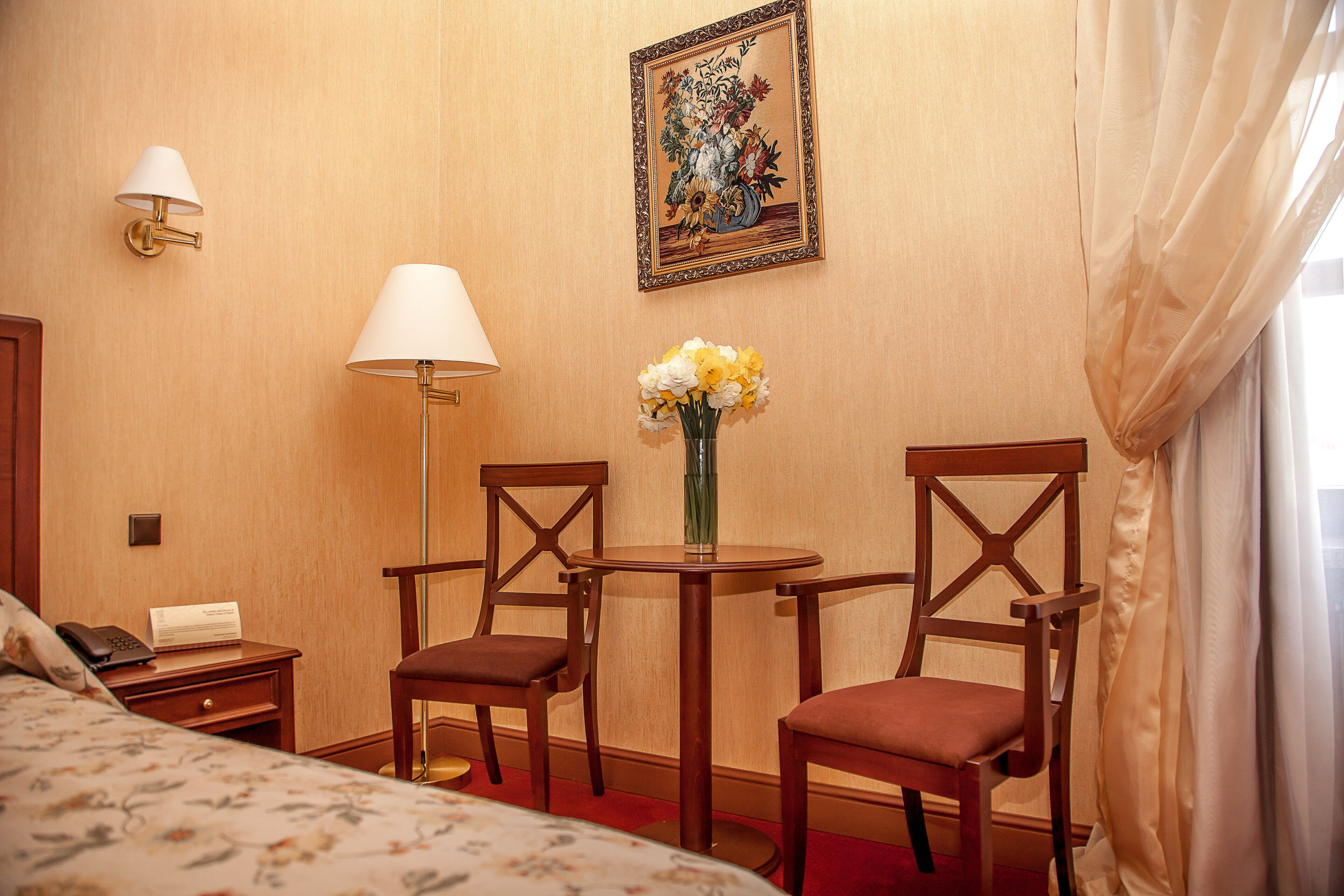 A hotel room with a bed, two wooden chairs, a small table with a vase of flowers, a lamp, and a painting on the wall.