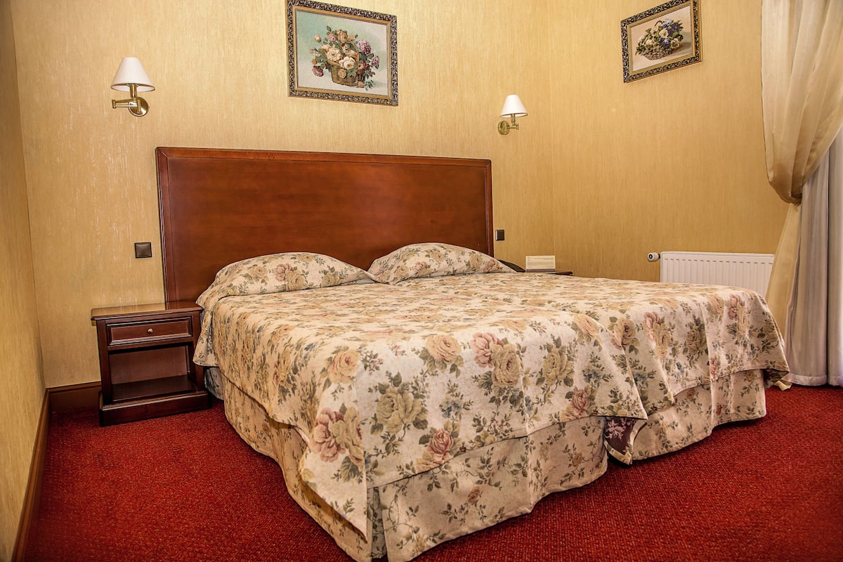 A hotel room with a wooden headboard, floral bedspread, two wall-mounted lamps, and a red carpet.