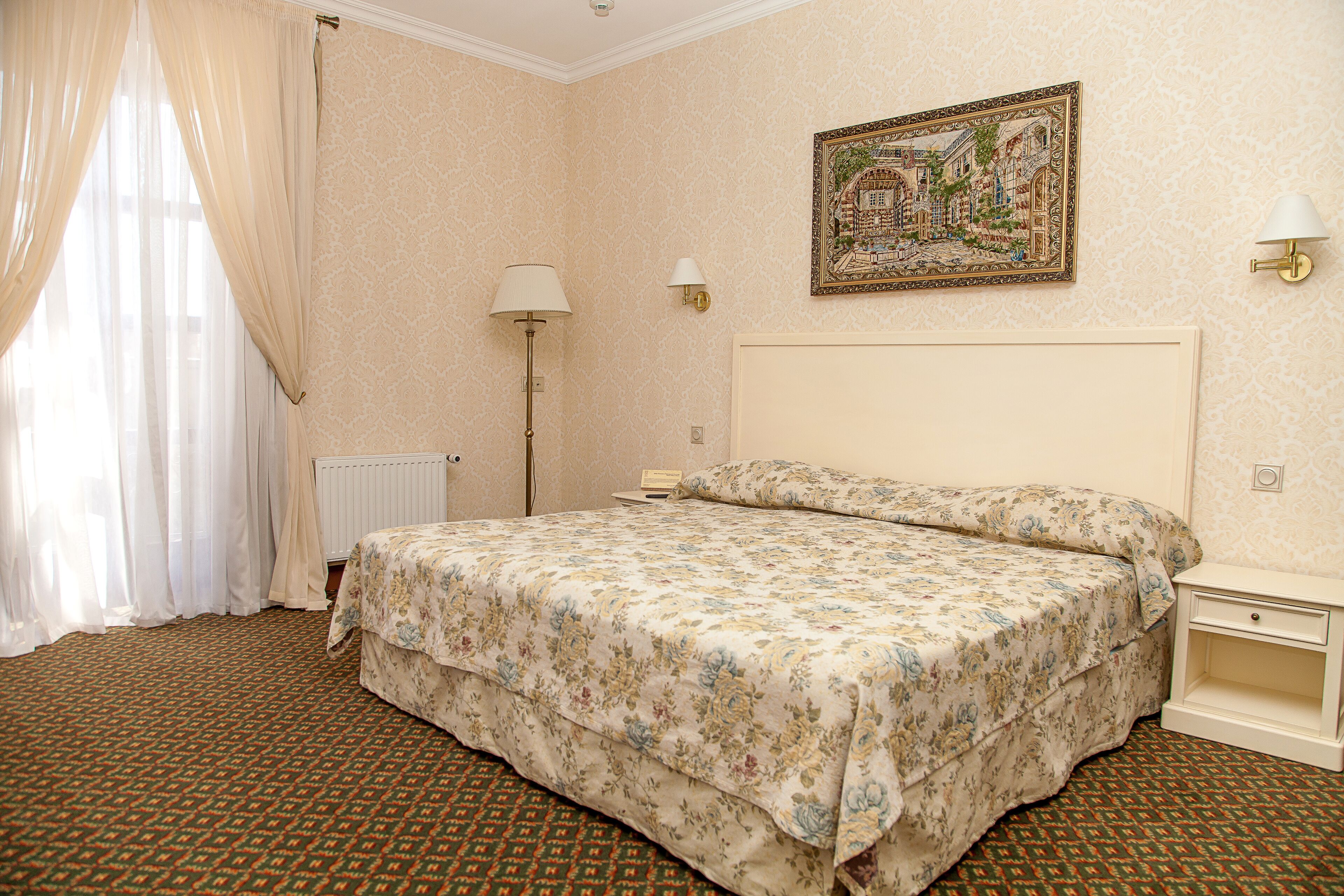 Una camera d'albergo con un letto, un comodino, una lampada, un termosifone e un quadro appeso al muro.