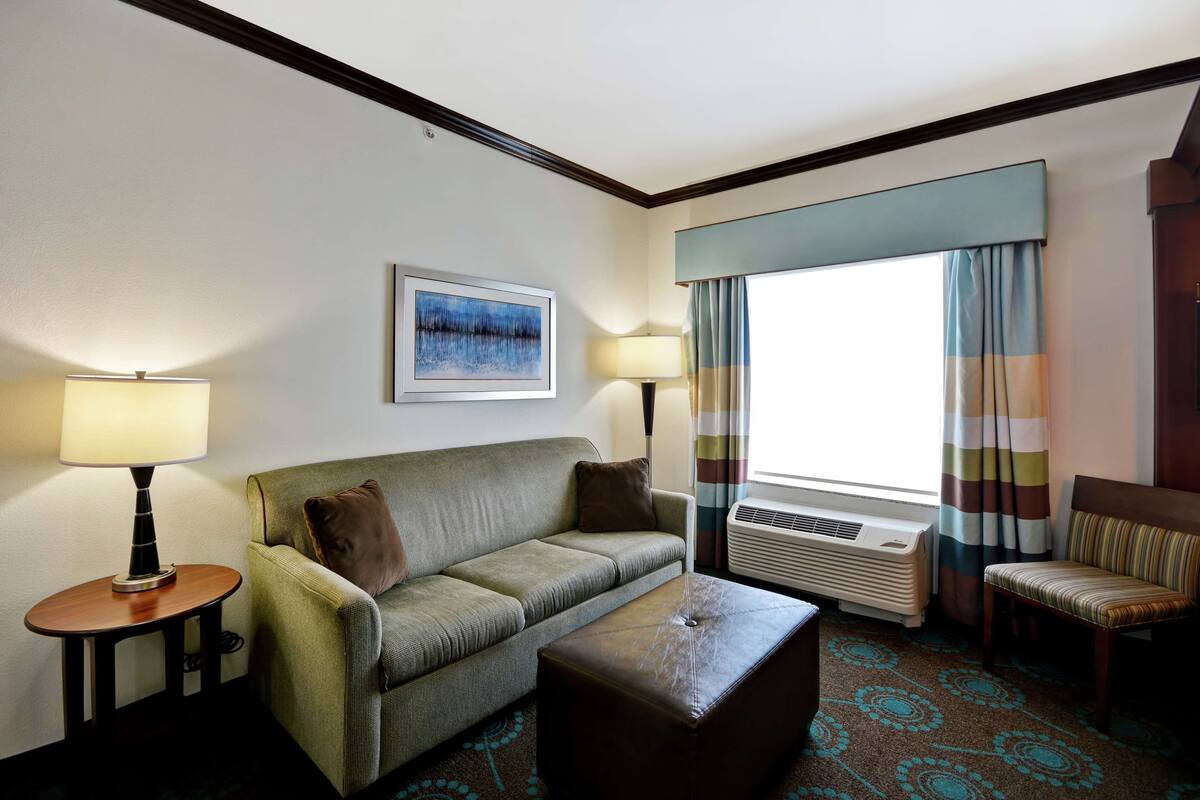 Ein Hotelzimmer mit einer Couch, einem Sessel, einem kleinen Tisch mit Lampe, einem Fenster mit Vorhängen und einer Wand-Klimaanlage.
