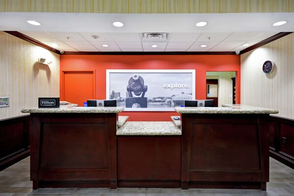 Un comptoir d’accueil d’hôtel avec une grande enseigne indiquant « Explorez ! », deux ordinateurs HP et une enseigne « Hilton Honors ».