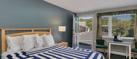 A hotel room with a bed, a desk, two chairs, a lamp, and a view of the street through the window.