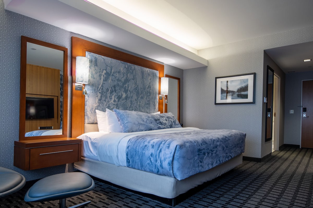 A hotel room with a large bed, a wooden nightstand, a mirror, a wall-mounted lamp, and a framed picture.