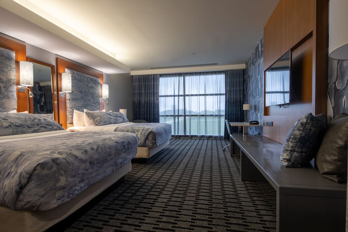 A modern hotel room with two beds, a large window, a flat-screen TV, and a desk.