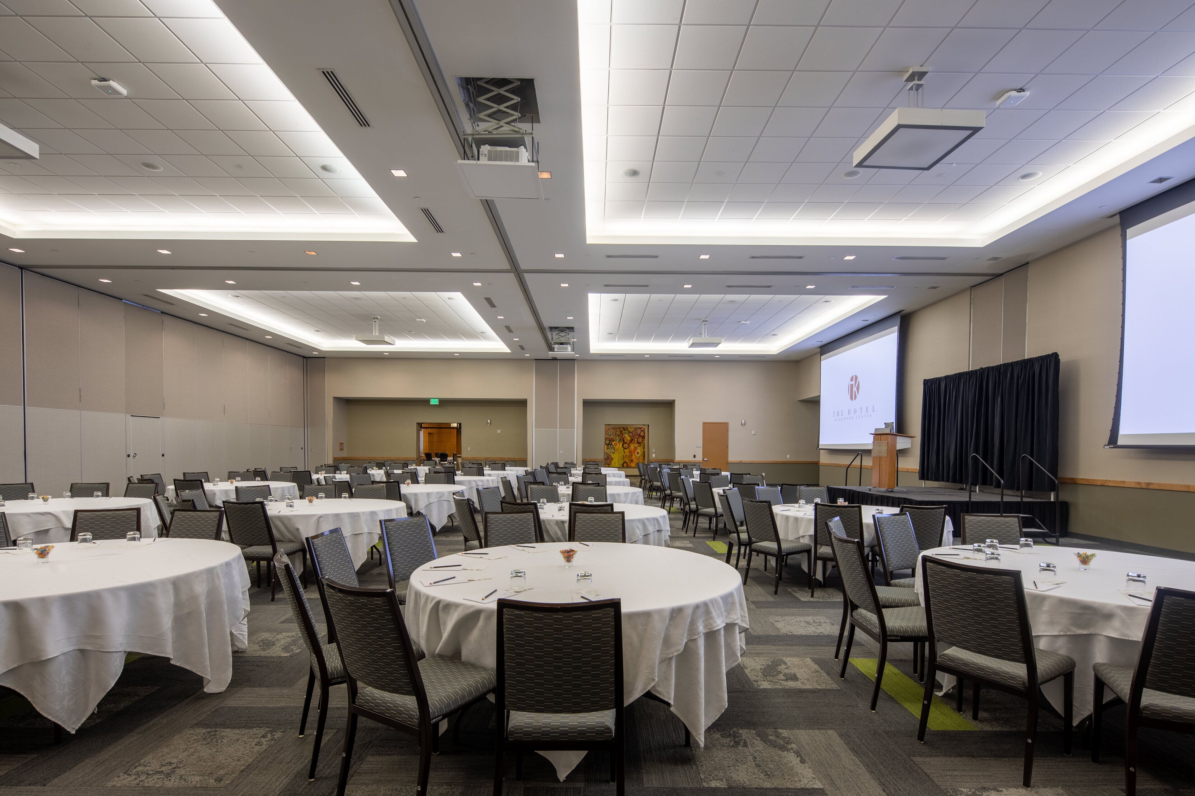 Un salón de conferencias con varias mesas redondas, sillas y una pantalla para presentaciones.