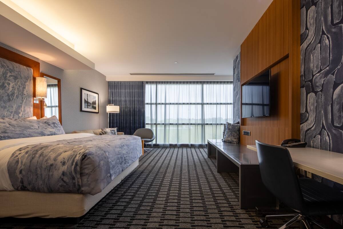 A hotel room with a large bed, a desk, a chair, a TV, and a view of the city through large windows.