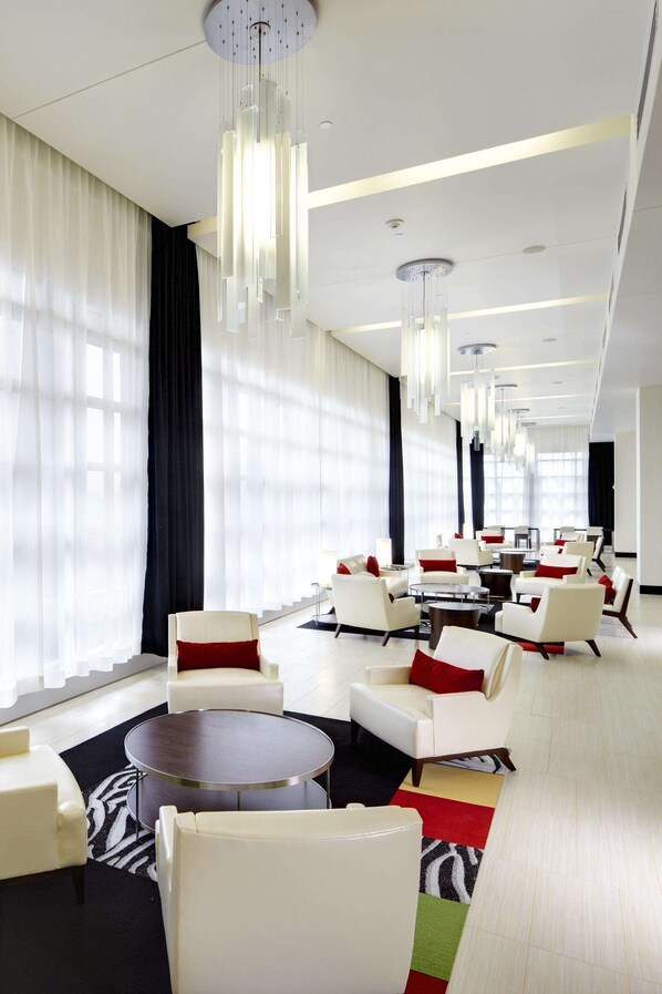 A modern hotel lobby with white and red upholstered chairs, a central round table, and a large window with sheer curtains.