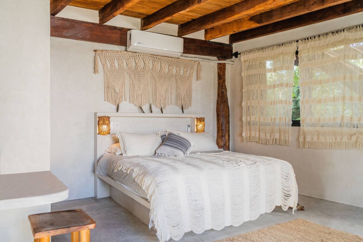 A bedroom with a bed, a wooden bench, a macrame wall hanging, and a window with curtains.