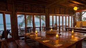 A wooden dining area with tables set for two, overlooking a beach with palm trees. - Amansala (Tulum)