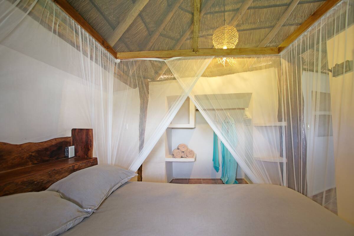 A bedroom with a wooden headboard, a bed with white linens, a nightstand, a small shelf with rolled towels, and a hanging light fixture.